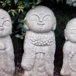 Three serene stone Buddha statues nestled in a tranquil garden, surrounded by lush greenery and vibrant flowers.